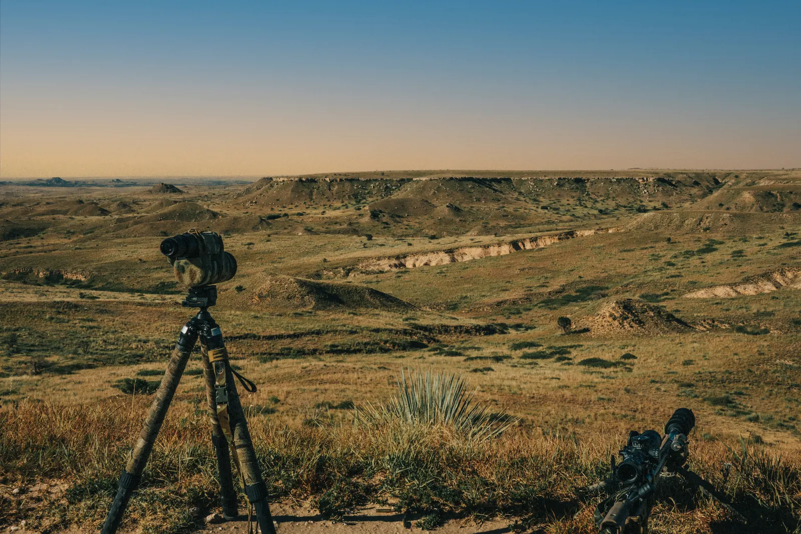 A spotting scope on a tripod and a rifle on its bipod at the edge of a ridge, looking out over wide prairie badlands under an evening sky.
